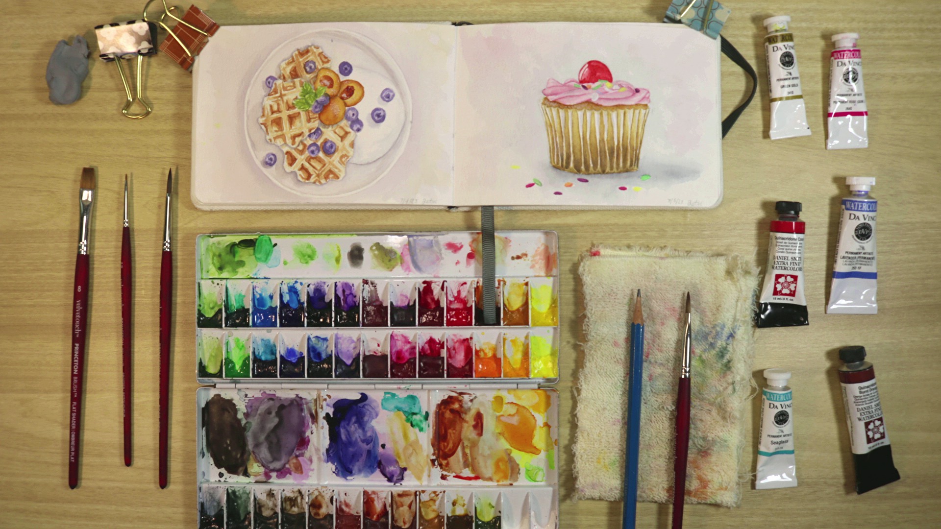 A sketchbook with paintings of desserts lays on a desk surrounded by watercolor supplies.