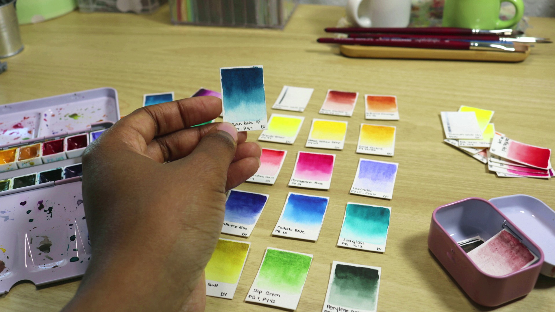 A hand holds a watercolor paint swatch and hovers over an arrangement of other paint swatches. A watercolor palette lays open to the left.