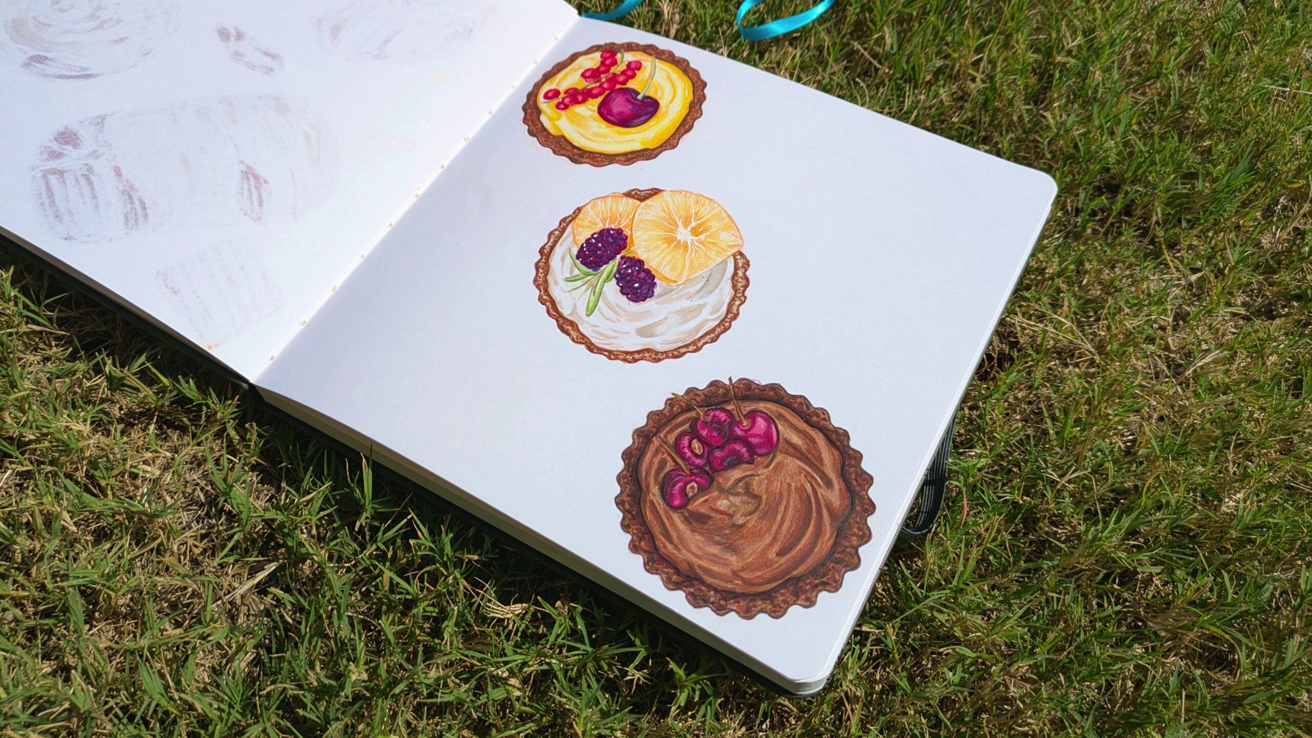 A sketchbook lays open on grass showcasing three different dessert tarts done with alcohol markers.