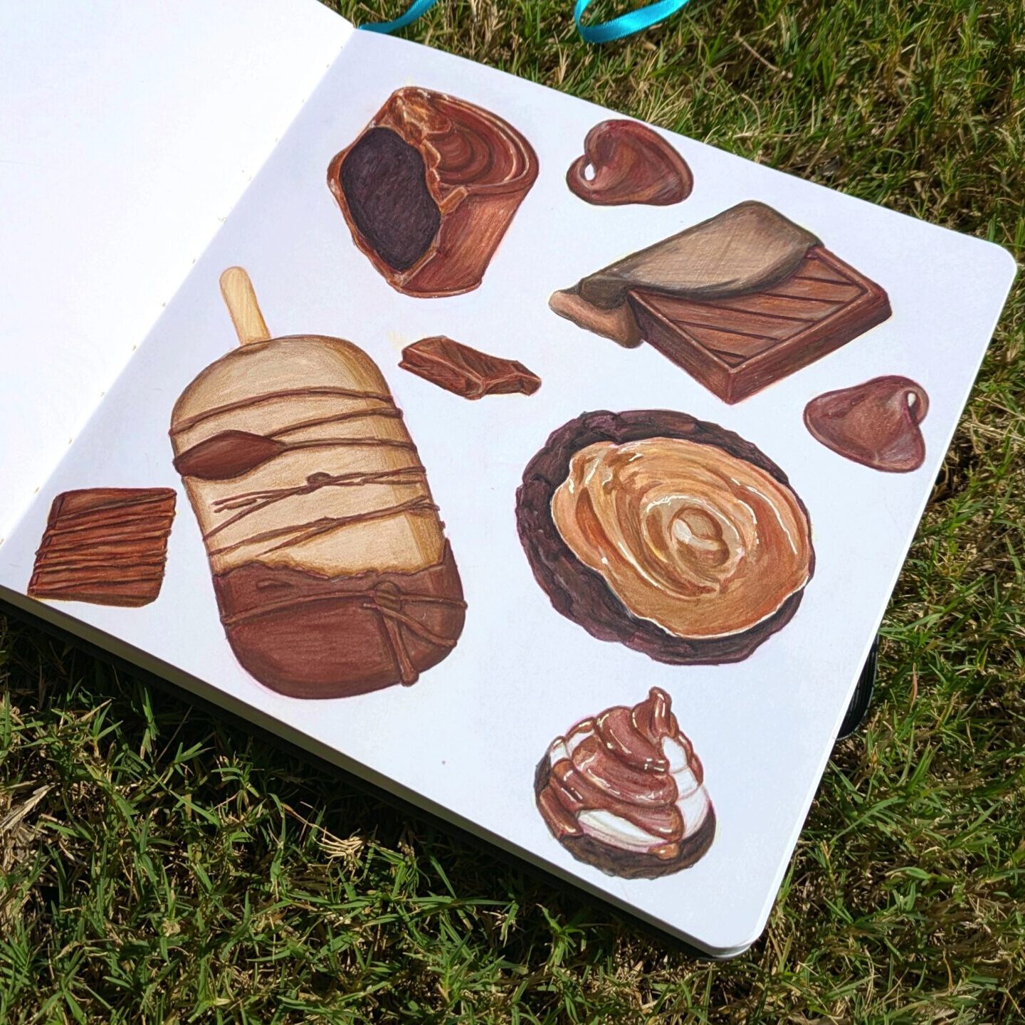 A sketchbook lays open on the grass. The page contains drawings of chocolate desserts: ice-cream bar, cookie, chocolate square, chocolate chips, and a cupcake